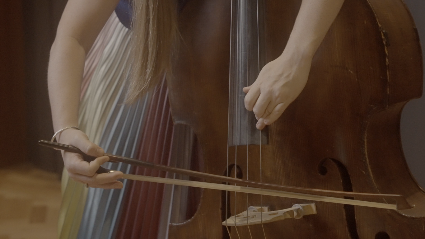 Close up of someone playing a double bass with a German style double bass bow. The person is wearing a multi coloured skirt.