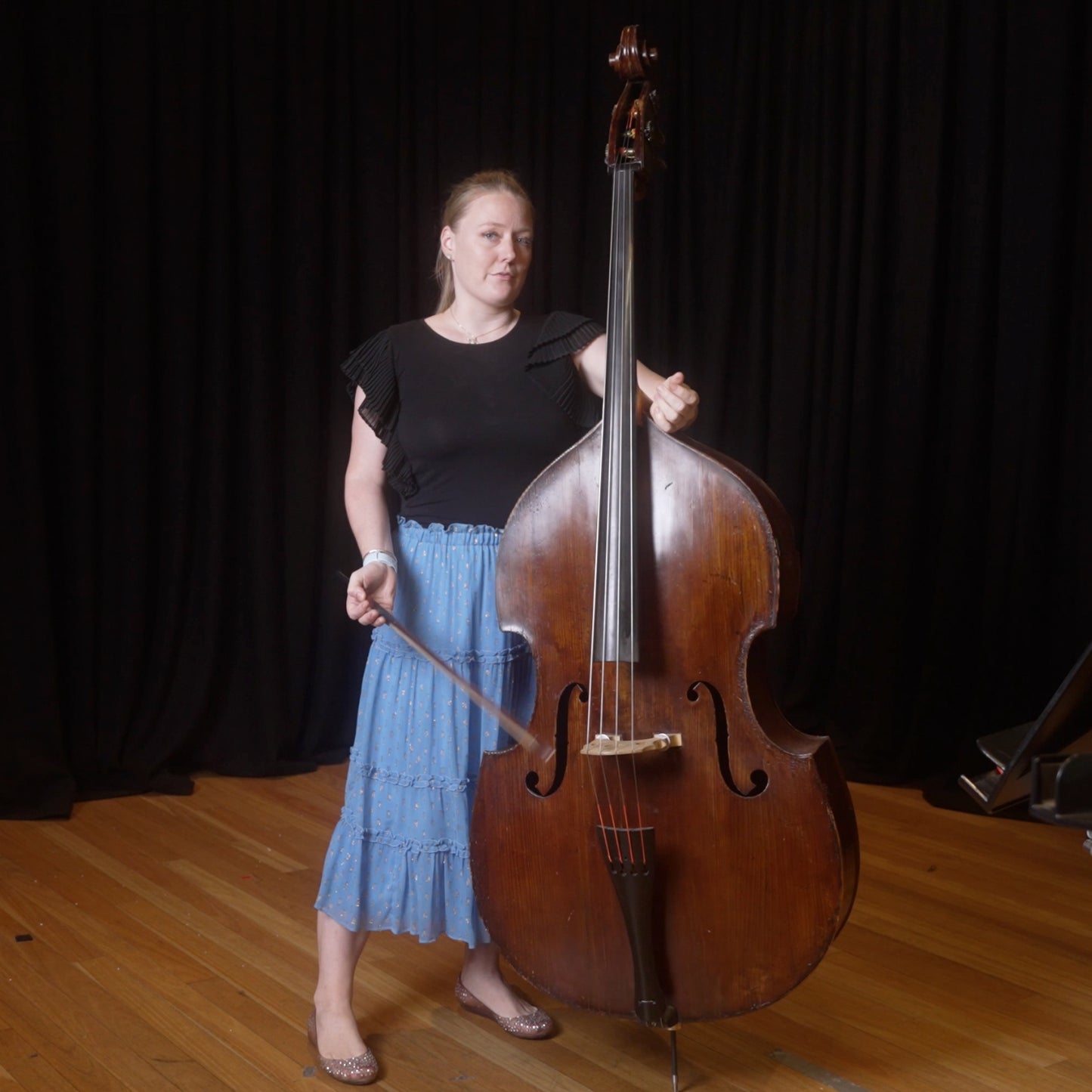 Phoebe Russell standing next to a double bass, holding a bow, with a black shirt and a blue skirt.